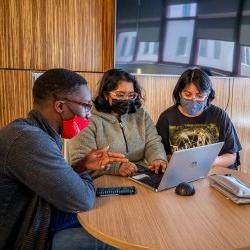 A group of students sit together at a table, looking at a laptop.